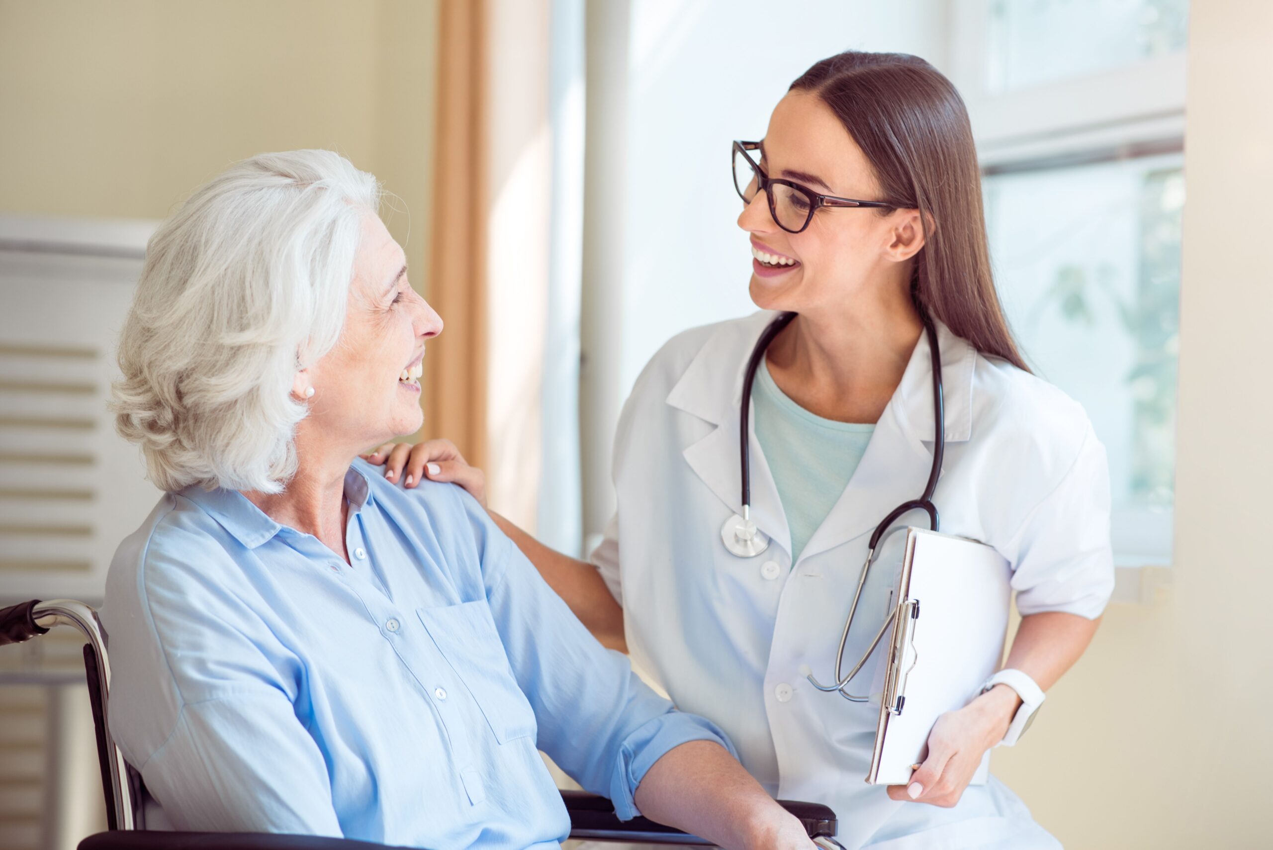 A smiling healthcare professional converses warmly with an elderly patient in a wheelchair, highlighting compassionate care in a medical setting.