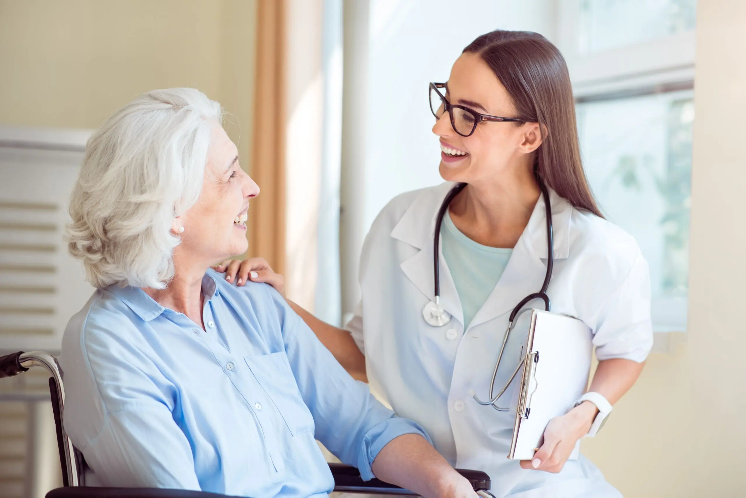 A smiling healthcare professional converses warmly with an elderly patient in a wheelchair, highlighting compassionate care in a medical setting.