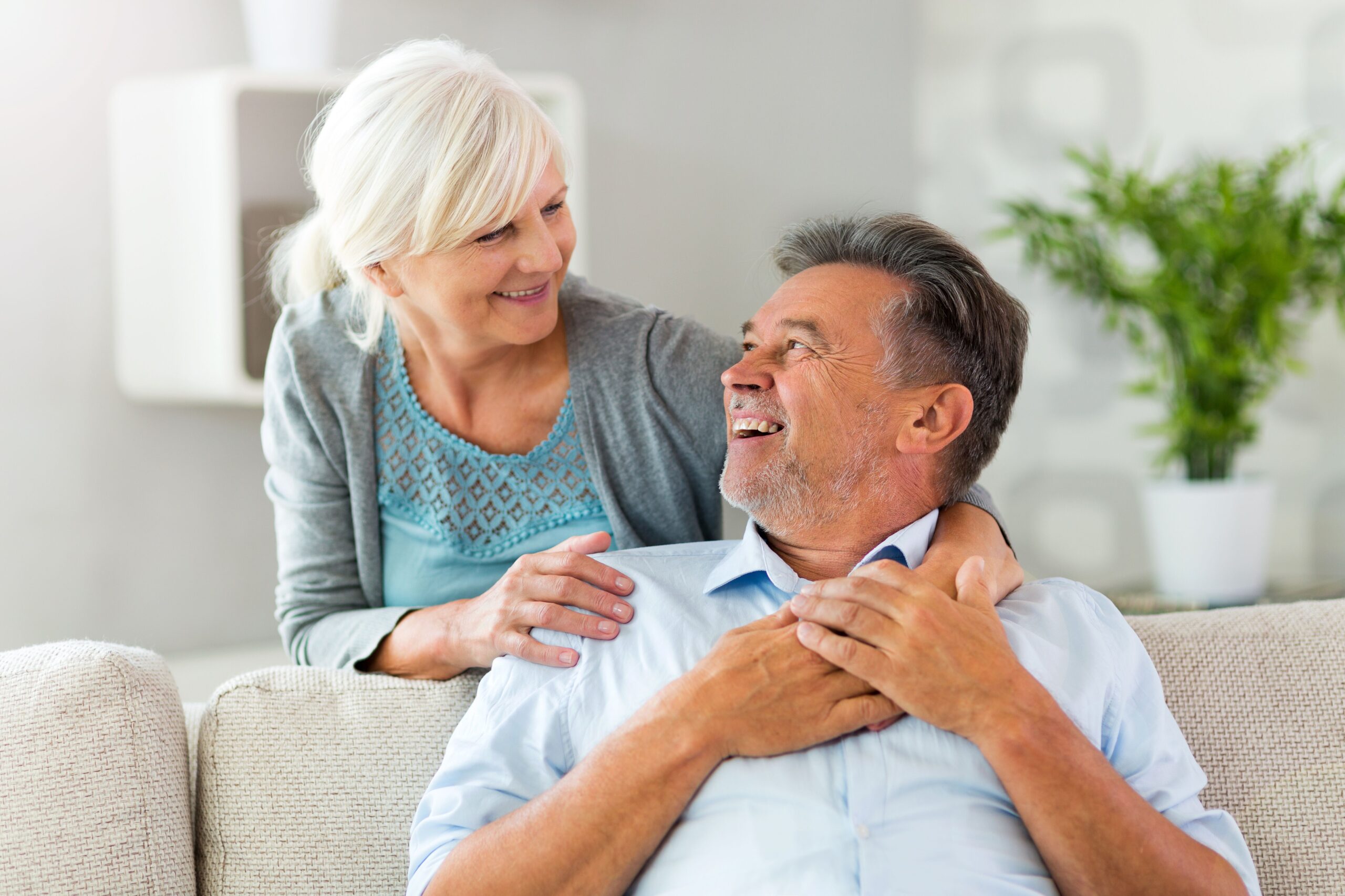 An elderly couple shares a joyful moment at home, smiling and looking at each other affectionately while seated on a sofa.
