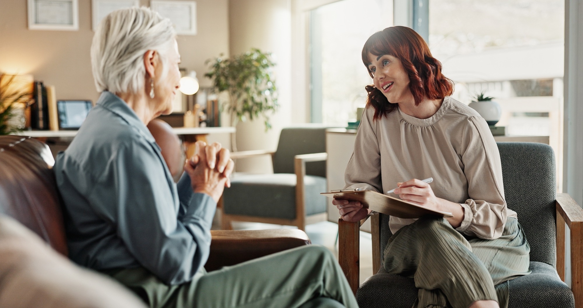 A therapist engages in a conversation with an older woman, taking notes in a cozy, well-lit office, highlighting a supportive counseling session.